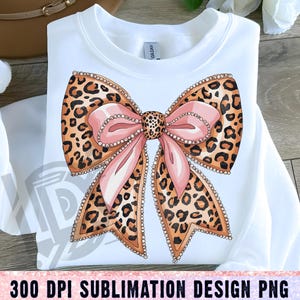 May include: A white shirt with a pink and brown leopard print bow with rhinestones. The bow is centered on the shirt.