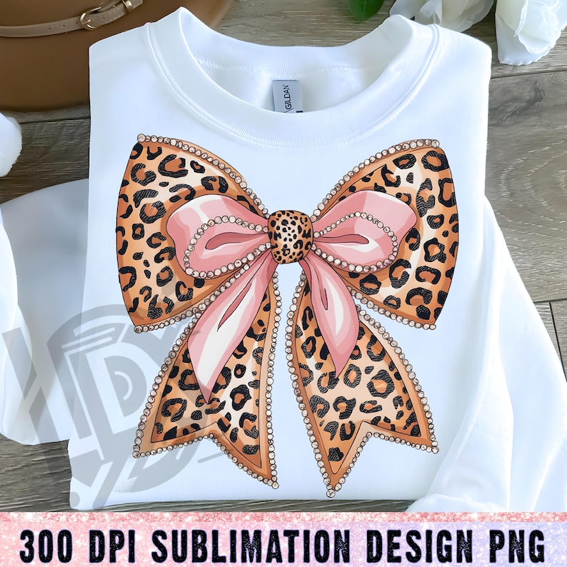 May include: A white shirt with a pink and brown leopard print bow with rhinestones. The bow is centered on the shirt.