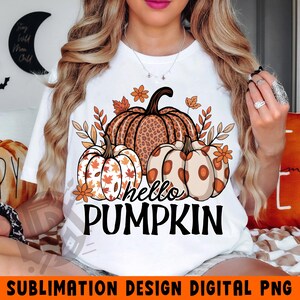 Könnte beinhalten: Weißes T-Shirt mit einem herbstlichen Design. Es zeigt drei Kürbisse in Braun-, Orange- und Cremetönen, umgeben von Blättern und Blumen. Der Text "hello PUMPKIN" ist schwarz. Digitales PNG-Design.