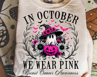 May include: A beige sweatshirt with a Halloween-themed graphic. The design features a ghost wearing a witch hat, surrounded by pink flowers, bats, and a pink pumpkin. The text reads "IN OCTOBER WE WEAR PINK Breast Cancer Awareness."