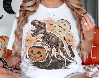 May include: White t-shirt featuring a Halloween-themed design. The graphic depicts a skeletal T-Rex holding a jack-o'-lantern, with a full moon and bats in the background. The text "SUBMLIMATION DESIGN DIGITAL PNG" is at the bottom.