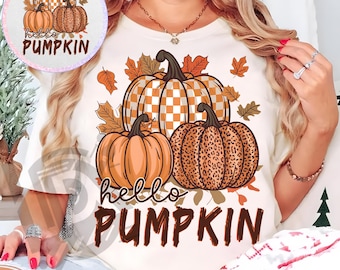 May include: Cream-colored t-shirt featuring a fall-themed pumpkin design. The design includes three pumpkins in orange, checkered, and leopard print patterns, with the words "hello PUMPKIN" in brown. Autumn leaves surround the pumpkins.