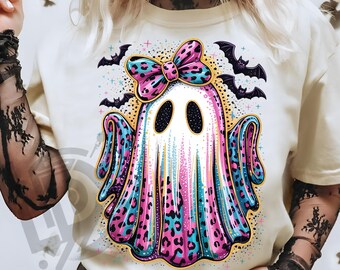 May include: A cream-colored t-shirt featuring a colorful ghost design with a bow, leopard print accents, and bats. The ghost is white with black eyes and a pink, blue, and purple leopard print design. The text "300 DPI SUBLIMATION DESIGN PNG" is at the bottom.