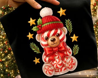 May include: Black t-shirt with a Christmas bear design. The bear has red and white candy cane stripes, a red knitted hat and bow. Gold stars and green sprigs surround the bear. The text "300 DPI SUBLIMATION DESIGN PNG" is at the bottom of the image.