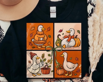 May include: Black sweatshirt featuring a four-panel design with cartoon ducks in autumn settings. Each panel shows a duck with fall elements like pumpkins, leaves, and a basket. The text "300 DPI SUBLIMATION DESIGN PNG" is at the bottom.