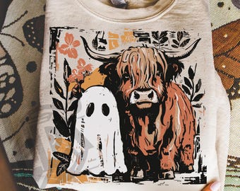 May include: A beige sweatshirt featuring a graphic of a Highland cow and a ghost. The cow is brown with large horns, and the ghost is white. Floral and leaf accents surround the cow and ghost. The text "300 DPI SUBLIMATION DESIGN PNG" is at the bottom.