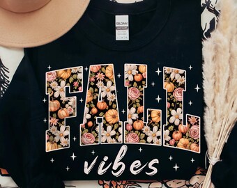 May include: Black sweatshirt with the words "FALL vibes" in a floral design with pumpkins and flowers. The sweatshirt has a crew neck and long sleeves. The design is in shades of orange, white, and pink.