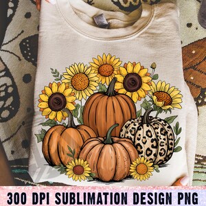 Peut inclure: Un sweat-shirt beige avec un motif d'automne. Le motif comprend des citrouilles oranges, des tournesols jaunes et des feuilles vertes. Une citrouille a un motif léopard. Le texte "300 DPI SUBLIMATION DESIGN PNG" est en bas.