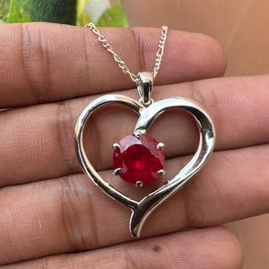 May include: A silver heart-shaped pendant necklace with a large, round, red gemstone in the center.