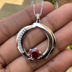 Peut inclure: Un collier avec un pendentif en argent, orné d'une pierre rouge au centre d'un cercle. Le cercle est entouré de petites pierres claires.