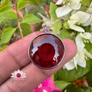 May include: A round, faceted, deep red gemstone pendant with a silver bezel. The pendant is held in a hand against a backdrop of green leaves and white flowers. The pendant has a loop at the top for a chain.