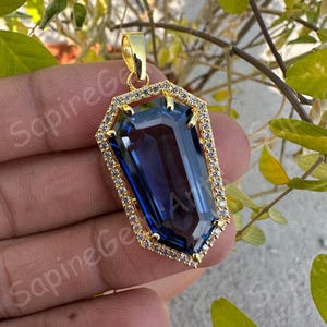 May include: A gold pendant featuring a large, rectangular, deep blue gemstone. The gemstone is surrounded by a frame of smaller, clear stones and a gold setting. The pendant is suspended from a gold bail.