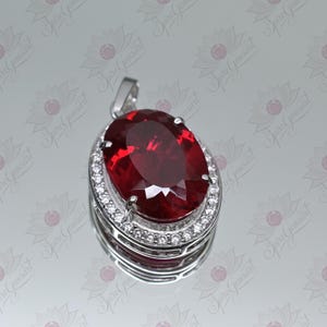 May include: A silver pendant featuring a large, oval-shaped, red gemstone set within a halo of smaller, white gemstones.