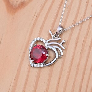 May include: A silver pendant necklace featuring a heart-shaped design. The pendant has a large, round, deep red gemstone at its center, surrounded by a halo of smaller, clear stones. The necklace chain is delicate and silver.
