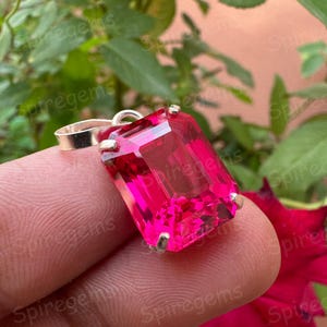 May include: A vibrant pink gemstone pendant with a rectangular cut. The gemstone is set in a silver metal setting with a loop for a chain. The background features green foliage and a red flower.
