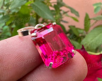 May include: A bright pink rectangular gemstone pendant. The gemstone is set in a silver metal setting with a loop for a chain. The background shows green leaves and a red flower.