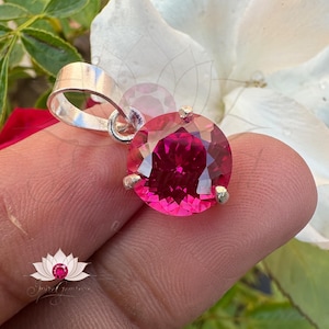 May include: A silver pendant featuring a large, round, faceted, pink gemstone. The pendant is resting on a finger.