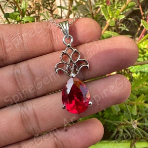 Könnte beinhalten: Eine silberne Halskette mit Anhänger, die einen tropfenförmigen, facettierten roten Edelstein aufweist. Der Anhänger hat ein verziertes, durchbrochenes Design über dem Edelstein. Die Halskette wird vor einem natürlichen grünen Hintergrund präsentiert.