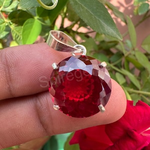 May include: A faceted, round, deep red gemstone pendant with a silver bail. The pendant is held up against a background of green leaves and a red rose. The word "Spireg" is visible on the gemstone.