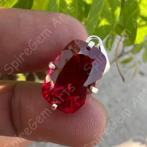 May include: A faceted, rectangular ruby gemstone pendant with a silver bail and prongs. The deep red stone is held in place by four silver prongs. The pendant is displayed against a blurred green background.