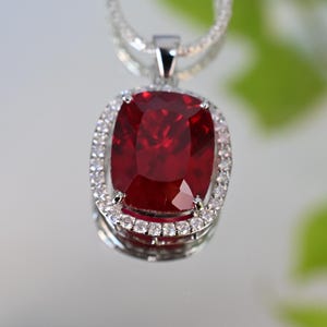Op de afbeelding: Een zilveren hangerketting met een grote, kussenvormig geslepen, dieprode edelsteen. De edelsteen is omgeven door een halo van kleine, heldere, sprankelende stenen. De ketting is ook zilver en heeft een delicaat, getextureerd ontwerp.