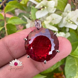 May include: A red gemstone pendant with a silver setting. The gemstone is round and has a faceted cut. The pendant is on a finger.