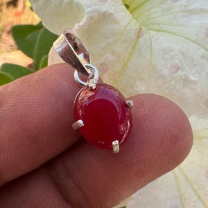 May include: A silver pendant with a large, oval-shaped, deep red gemstone. The gemstone is held in place by four silver prongs. The pendant has a silver loop at the top for a chain. The background is a blurred white flower.