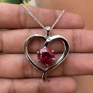 May include: A silver heart-shaped pendant necklace with a large, round, dark pink gemstone in the center.