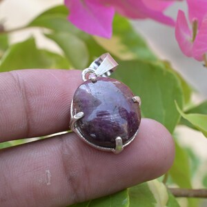 May include: A silver pendant featuring a large, oval-shaped, deep burgundy gemstone. The stone is set in a simple silver bezel with a small loop for a chain. The background includes green leaves and pink flowers.