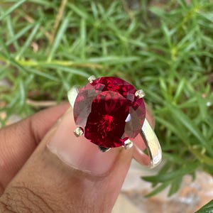 May include: A silver ring featuring a large, faceted, round, deep red gemstone. The gemstone is held in place by four prongs. The ring is set against a background of green foliage.