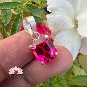 May include: A silver pendant featuring a large, bright pink, emerald-cut gemstone. The pendant is held in a hand against a blurred background of green leaves and white flowers.