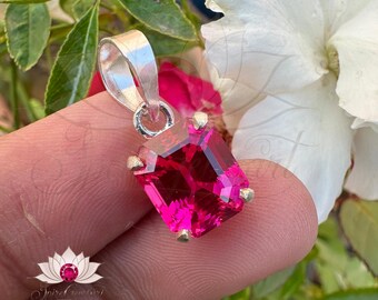 May include: A silver pendant featuring a large, bright pink, emerald-cut gemstone. The pendant is held in a hand against a blurred background of green leaves and white flowers.