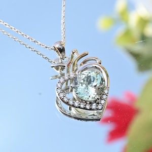 May include: A silver pendant necklace featuring a heart-shaped design. The pendant has a large, light green gemstone at its center, surrounded by smaller, clear stones. The necklace chain is delicate and silver.