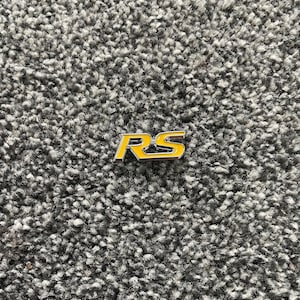 May include: A yellow and silver RS emblem, likely for a vehicle, rests on a gray, textured surface. The emblem features the letters "RS" in a bold, stylized font, with a yellow background and silver trim.