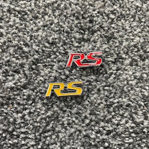 May include: Two metal "RS" emblems, one red and one yellow, on a grey carpet.