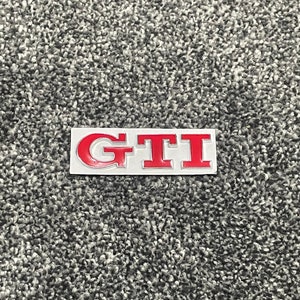 May include: A red "GTI" badge on a silver background. The badge is a popular car emblem.