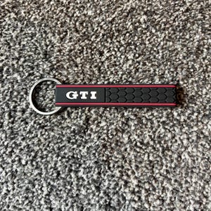 May include: Black and red GTI keychain with a silver key ring. The keychain has the letters GTI in white. The design includes a textured black pattern and a red border.