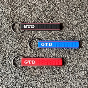 Może przedstawiać: Trzy breloczki do kluczy z białymi literami "GTD". Jeden jest czerwony z czarnym wzorem plastra miodu, drugi niebieski ze wzorem plastra miodu, a ostatni czarny z czerwonym wzorem plastra miodu. Każdy breloczek ma srebrny pierścień.