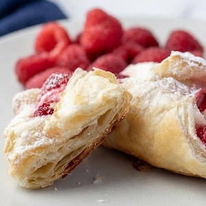 Peut inclure: Gros plan sur une assiette avec deux pâtisseries et des framboises fraîches. Les pâtisseries sont dorées, feuilletées et garnies de framboises rouges. Du sucre glace saupoudre légèrement les pâtisseries. Un tas de framboises rouges fraîches se trouve derrière les pâtisseries.