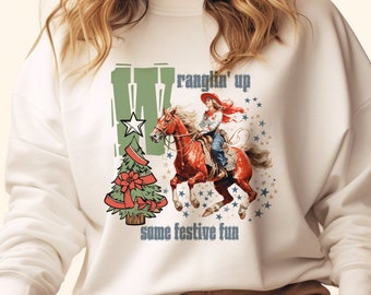 Puede incluir: Sudadera blanca con un diseño navideño que presenta a una vaquera montando un caballo con el texto "wranglin' up some festive fun".