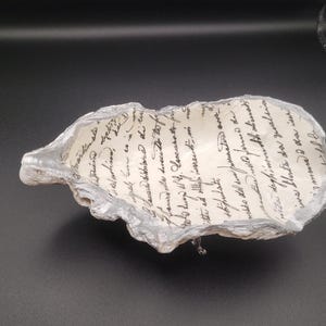 May include: A decorative oyster shell dish featuring a cream-colored interior with black cursive script handwriting. The exterior of the shell has a textured, metallic silver finish along the irregular, wavy edges, creating a unique bowl shape.