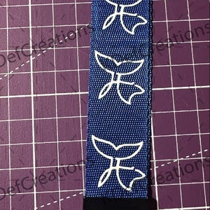 Puede incluir: Un cordón azul con diseños de cola de ballena blancos. El cordón tiene un clip negro y un lazo negro. El cordón mide aproximadamente 2,5 cm de ancho y 18 cm de largo.