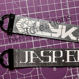 May include: Two keychains with black hardware and fabric straps. One strap is gray with the word "LYKN" in white, and the other is black with "JASPER" in white. The keychains are on a purple cutting mat.