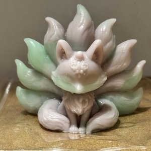 May include: A molded figurine of a nine-tailed fox with a smooth, semi-translucent finish. The creature features a light lavender body with pale mint green accents on the tips of its multiple tails and ears, and intricate embossed floral details.