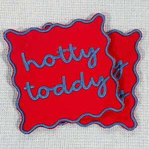 May include: Two red coasters with a wavy edge and blue embroidered trim. The words "hotty toddy" are embroidered in blue script on the coasters. The coasters are on a light gray surface.