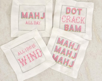 May include: Four white square coasters with embroidered text in pink and green. The coasters feature phrases like "MAHJ ALL DAY" and "DOT CRACK BAM". Each coaster has a dotted border.