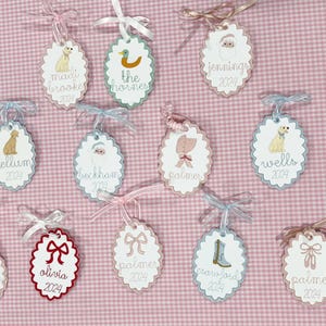 Può includere: Etichette regalo ovali con bordi smerlati, con illustrazioni e nomi come "Madi Brooke 2024" e "The Hornes". Ogni etichetta è adornata con un nastro e un fiocco, su uno sfondo rosa a quadretti.