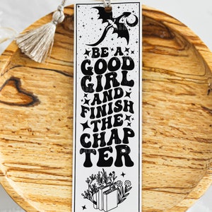 Puede incluir: Un marcapáginas blanco con un diseño de dragón negro y el texto "Be a good girl and finish the chapter". El marcapáginas tiene un borla y está sobre una superficie de madera.