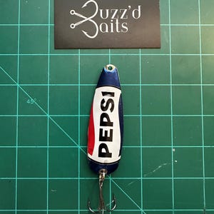May include: A blue, red, and white fishing lure with the word "PEPSI" printed on it. The lure has a treble hook and is on a green cutting mat.