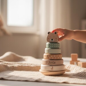 Peut inclure: Jeu d'empilage en bois avec une tête d'ours et des anneaux colorés. Les anneaux sont dans les tons de vert, beige et blanc. Le nom "Jonte" est imprimé sur l'un des anneaux. Une main d'enfant atteint la pièce du haut.